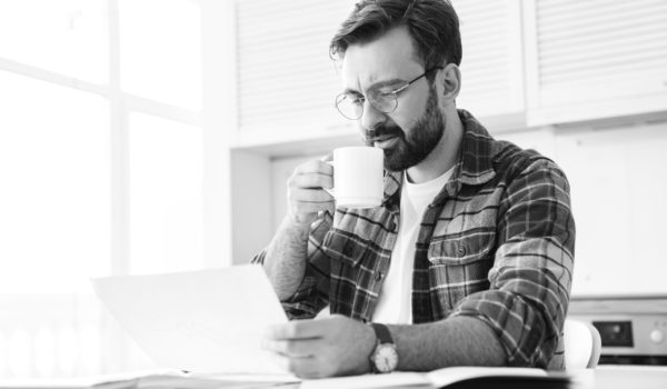Portrait of young bearded man wearing eyeglasses drinking tea while working with notes at home
